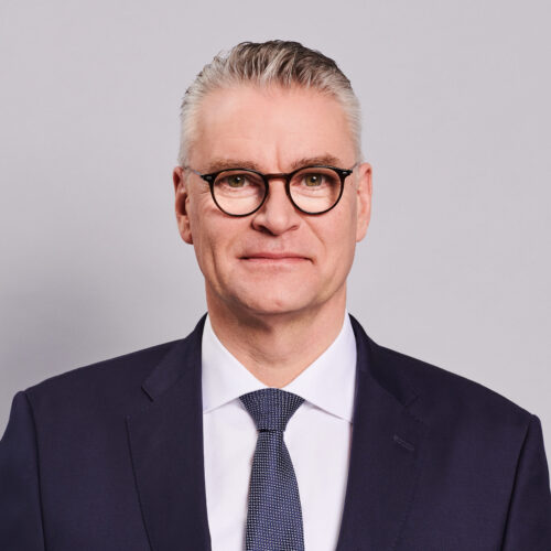 Portraitfoto von Dr. Constantin H. Alsheimer. Herr Alsheimer hat kurze graue Haare, keinen Bart und trägt Brille. Er trägt ein weißes Hemd, dunkles Jackett, Krawatte.