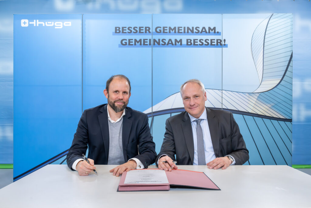 zwei Herren in Anzügen sitzen an einem Tisch. Links ist Dr. Tilman von Schroeter, rechts Dr. Matthias Cord zu sehen. Beide halten einen Stift. Vor ihnen auf dem Tisch liegt eine Unterschriftenmappe mit dem unterzeichneten Rahmenabkommen.